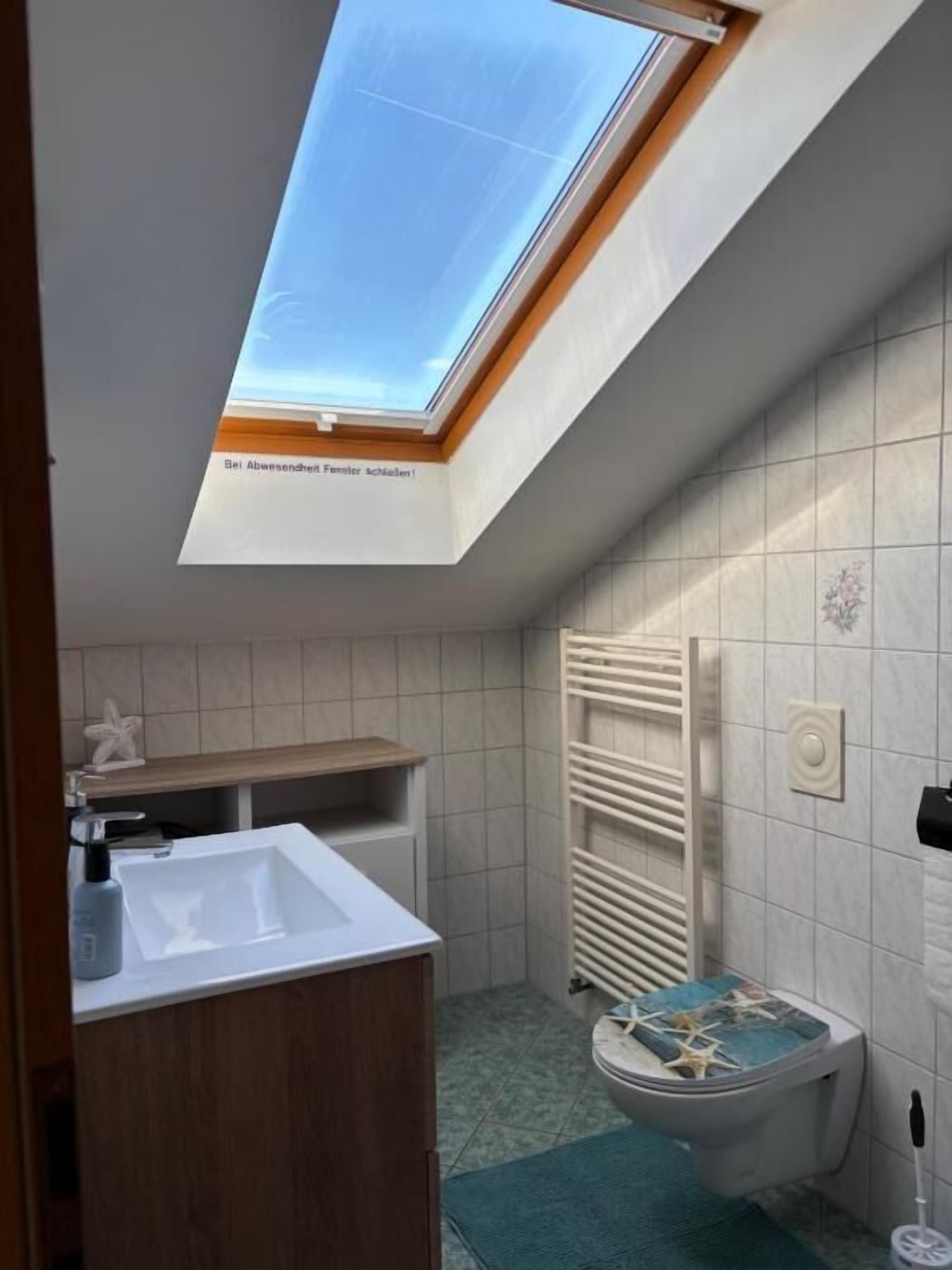 Ein modernes Badezimmer mit Oberlicht, einem weißen Waschbecken, einem Holz-Waschtisch, einer Toilette mit dekorativem Deckel und einem Handtuchhalter.