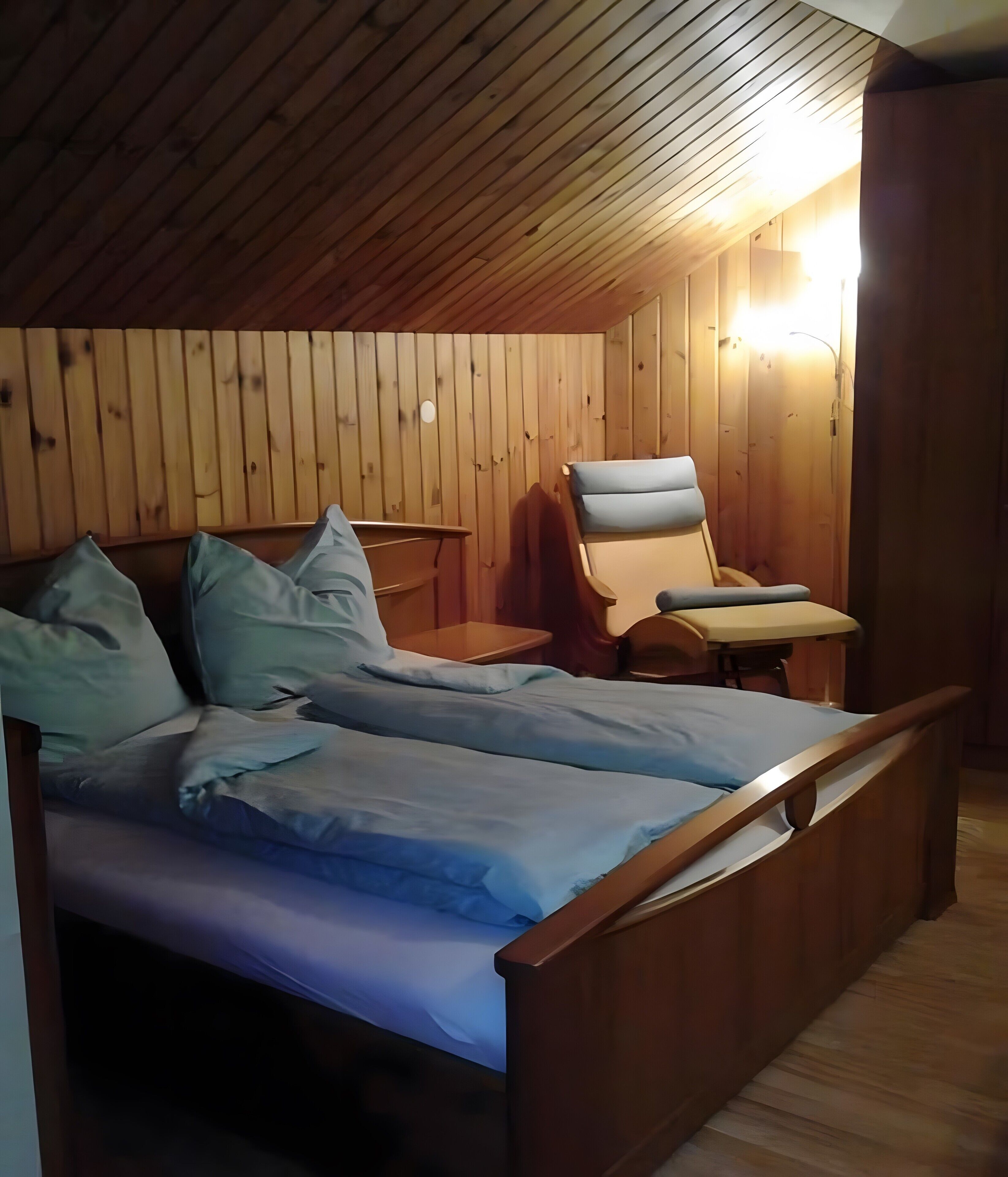 A room with a wooden ceiling and walls, a bed with blue bedding, a chair, and a small table.