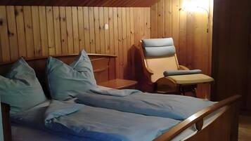 A room with a wooden ceiling and walls, a bed with blue bedding, a chair, and a small table.