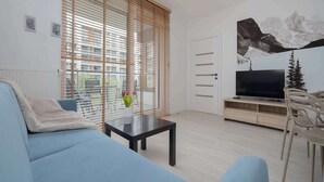 Ein modernes Wohnzimmer mit einer Couch, einem Couchtisch mit einer Vase, einem Fernseher auf einem Holzboden und großen Fenstern mit Jalousien.