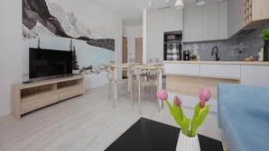 Ein modernes Wohnzimmer mit Flachbildfernseher, Holzesstisch, Stühlen und einer Küchenzeile mit Schränken und Pendelleuchten.