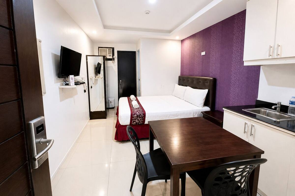 A hotel room with a bed, a desk, a chair, a television, a wardrobe, and a kitchenette.