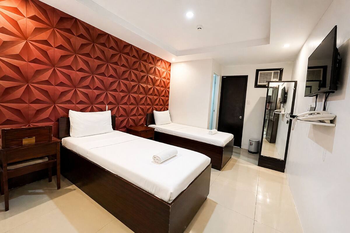 A hotel room with two beds, a wooden nightstand, a mirror, a television, and a wall with a geometric pattern.
