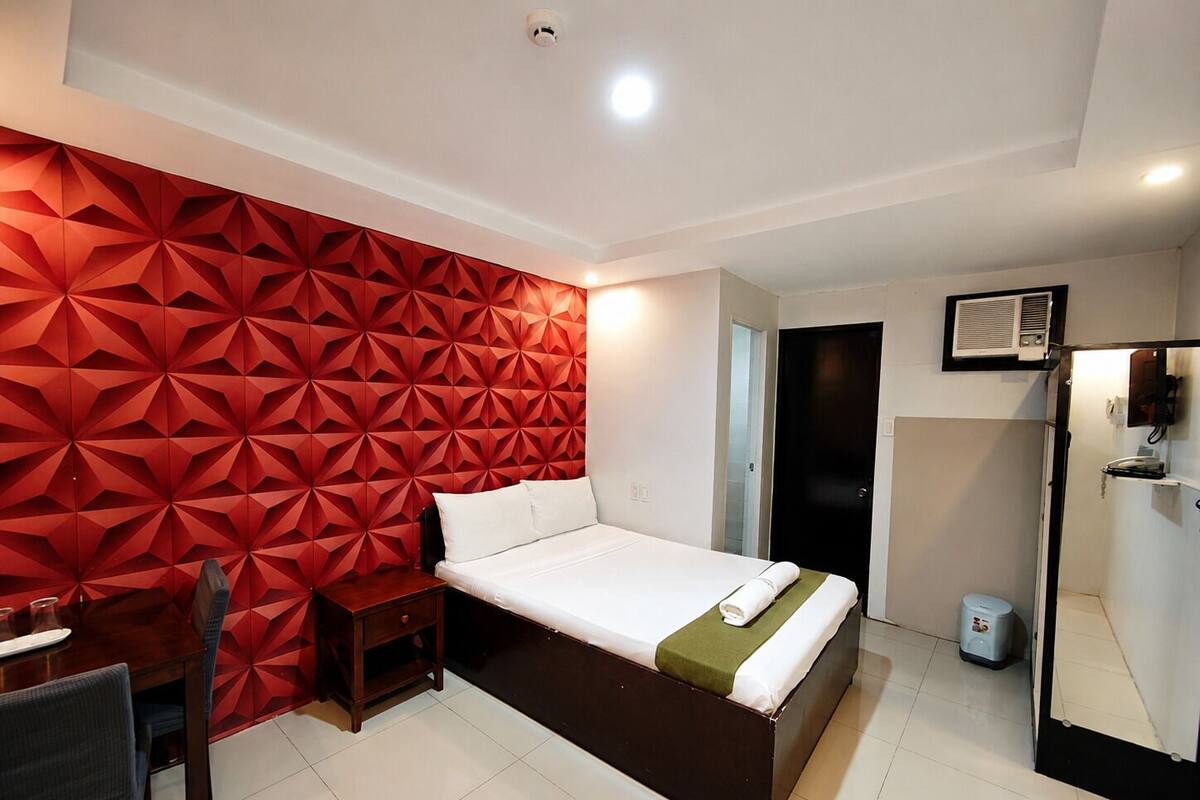 A hotel room with a bed, a desk, a chair, a red geometric wall, a black door, and a white tiled floor.