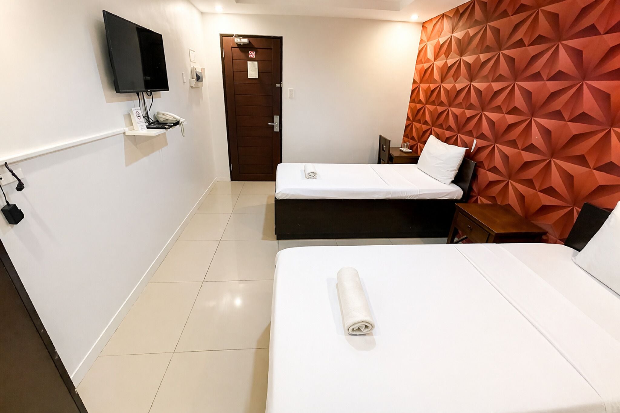 A hotel room with two beds, a TV mounted on the wall, a door, and a geometric-patterned wall.