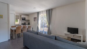 A modern living room with a grey sofa, a dining area with wicker chairs, a kitchen with white cabinets, and a flat-screen TV on a white stand. - Lacanau Stay, 100m to Lake & Pool (Lacanau)