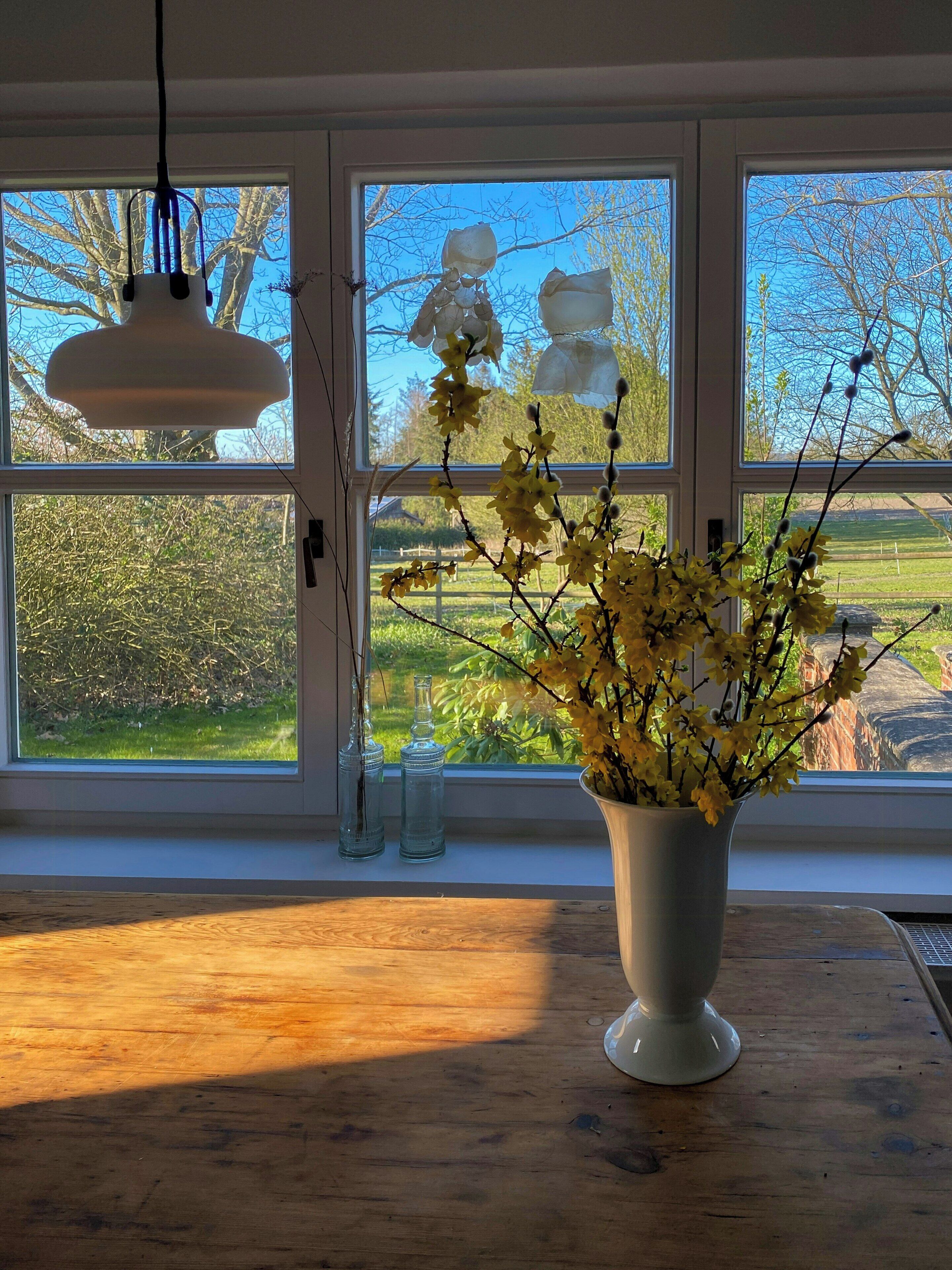 A room with a large window, a vase with yellow flowers, and a hanging light fixture.