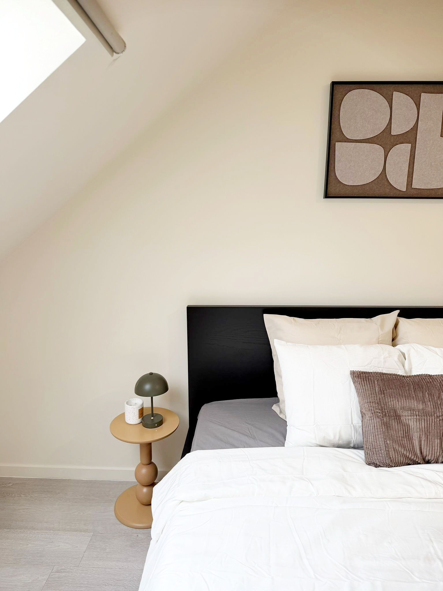A bedroom with a bed, pillows, a round table with a lamp, a wall art piece, and a sloped ceiling.