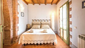 A bedroom with a wooden ceiling, a brick wall, a bed with a white bedspread, and a window with green shutters. - Villa with private pool among Siena's olive groves (Rapolano Terme)