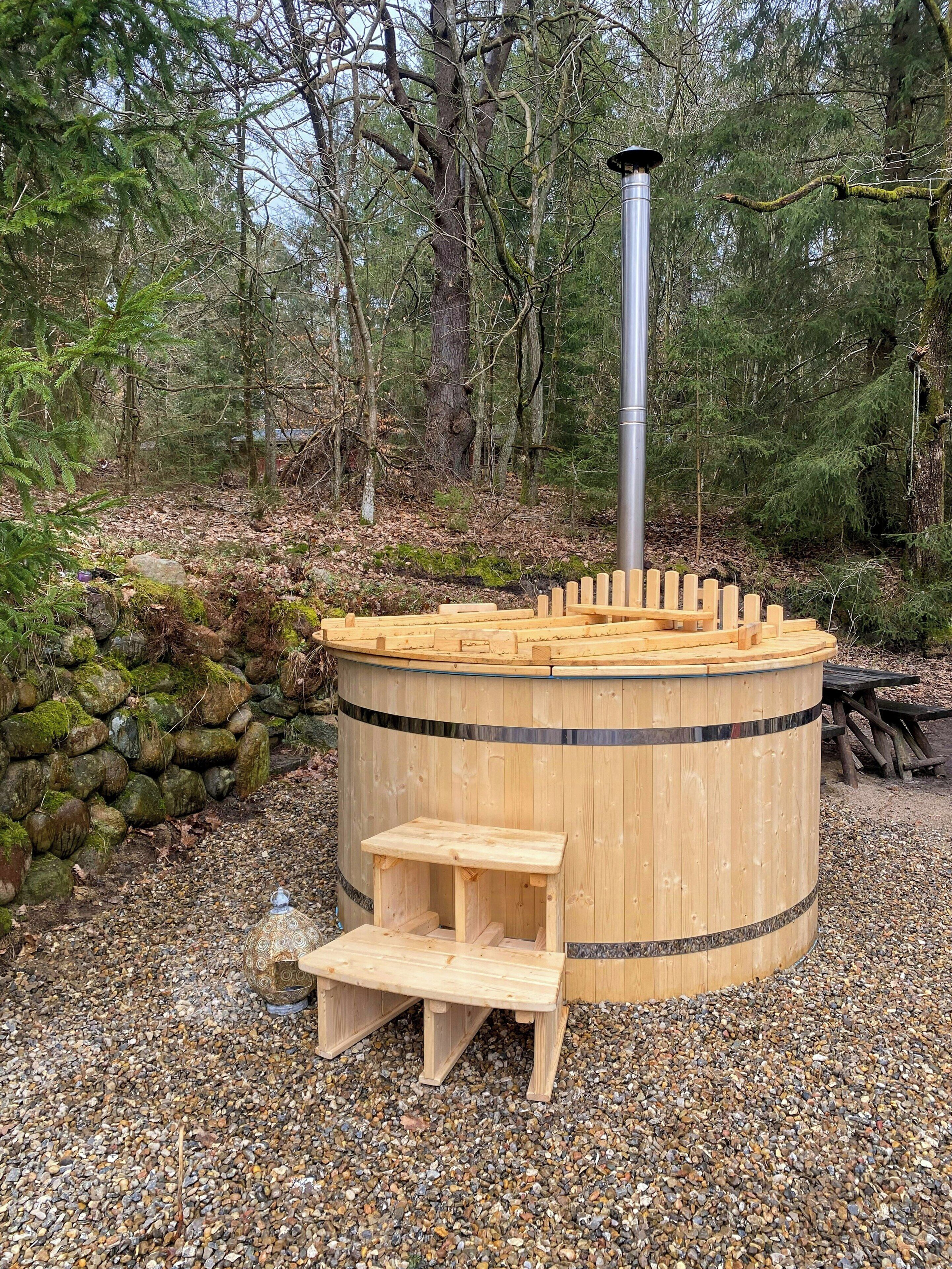 A wooden hot tub with a bench, surrounded by pebbles and trees.
