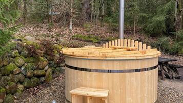 A wooden hot tub with a bench, surrounded by pebbles and trees.