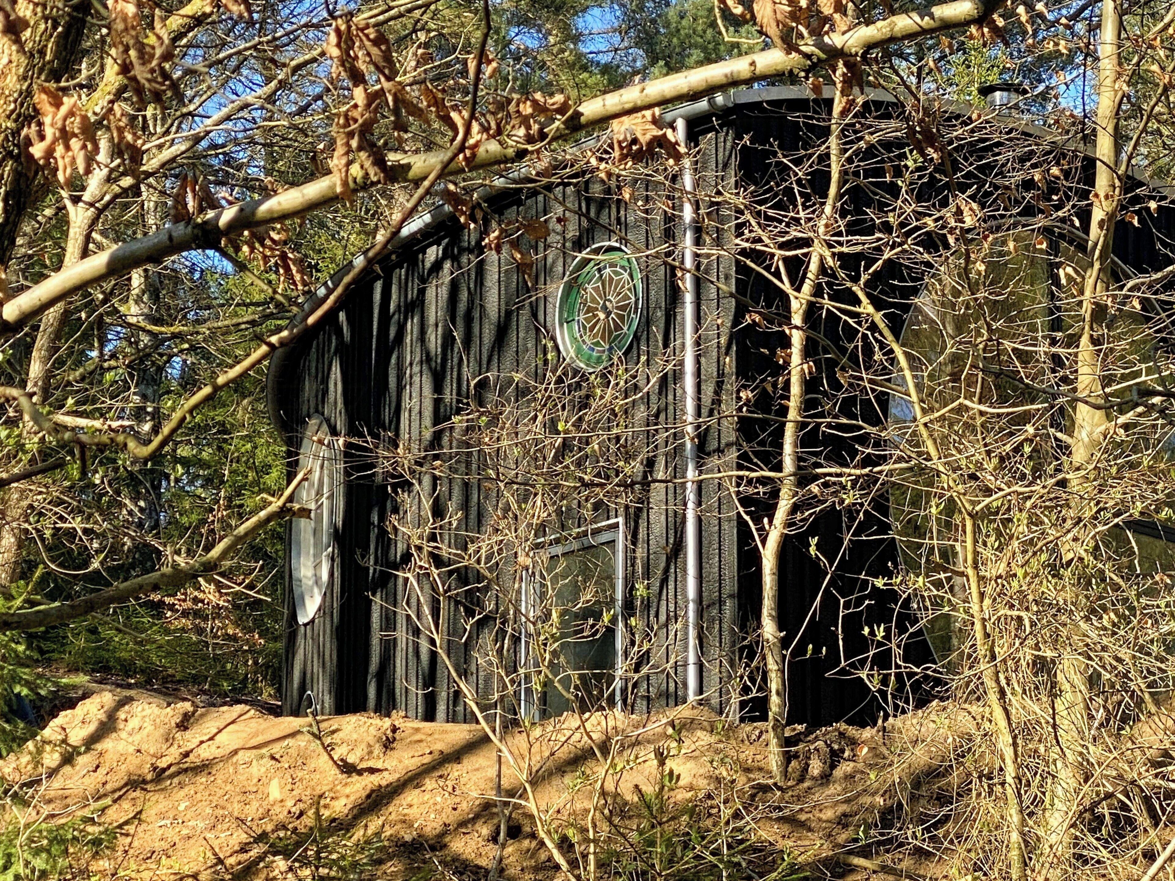 Une grange rustique, ornée d’un emblème circulaire sur la porte, entourée d’arbres sans feuilles et d’un sol en terre battue.