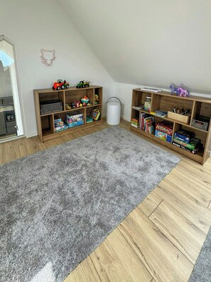 Una habitación con dos estanterías de madera llenas de juguetes y libros, alfombra gris y suelo de madera.