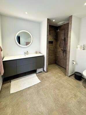 Un baño moderno con un espejo redondo, un lavabo blanco, un mueble de tocador oscuro, una ducha y un inodoro.
