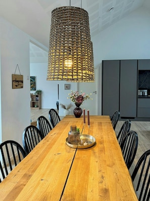 A modern dining area with a wooden table, black chairs, a hanging woven light fixture, and a vase with flowers.