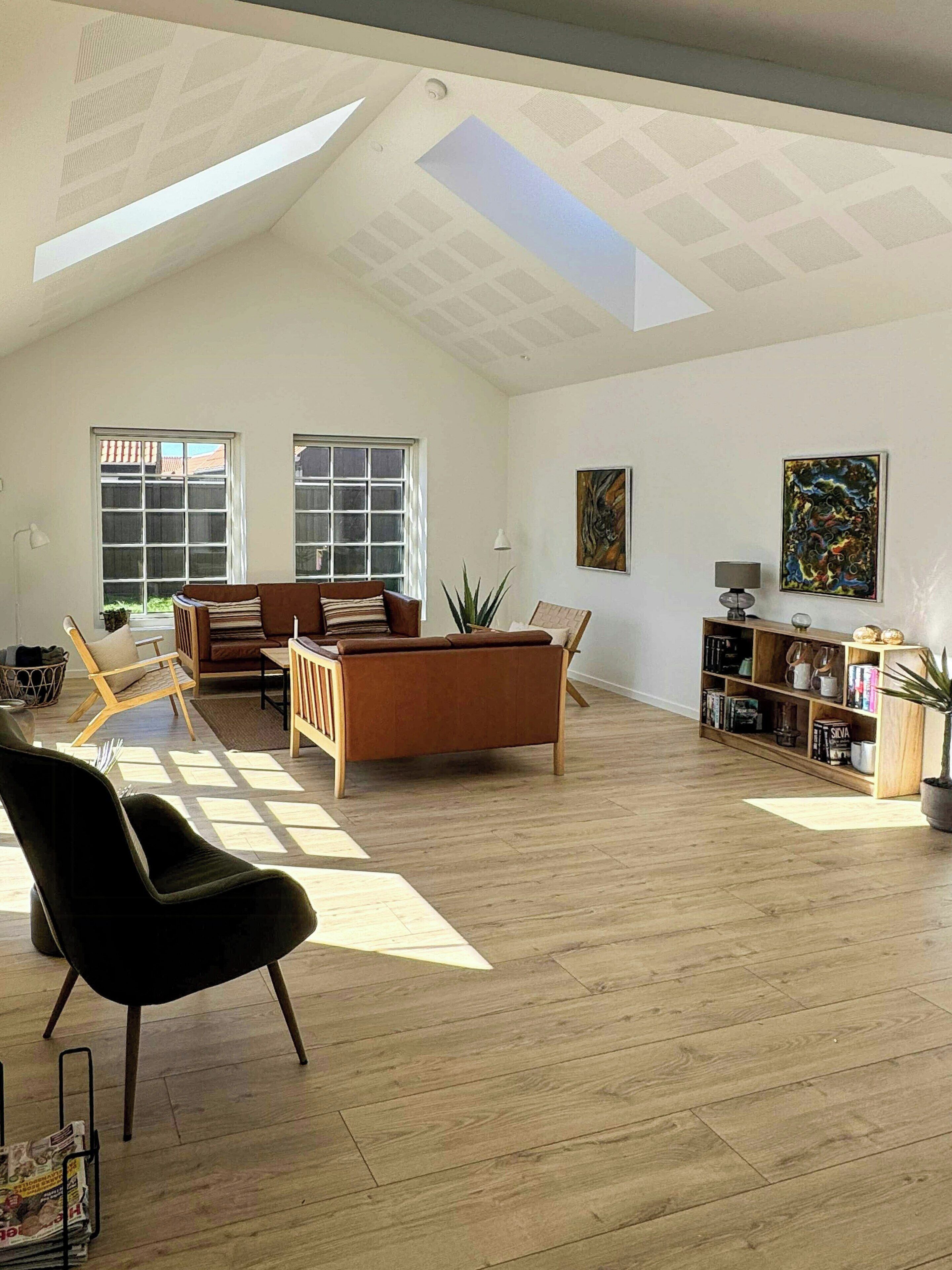 A spacious living room with a high ceiling, large windows, a brown leather sofa, a black chair, and a wooden bookshelf.
