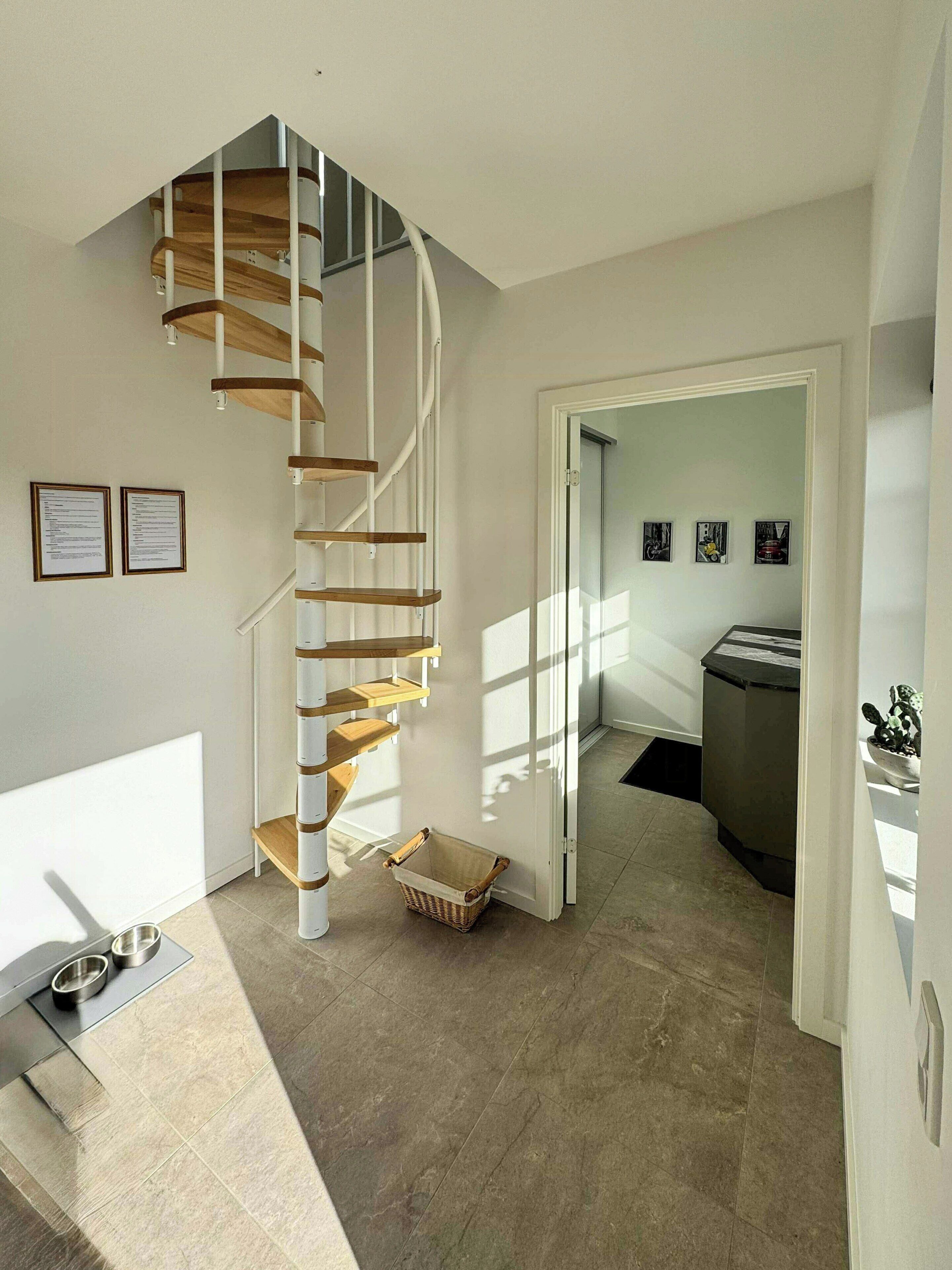 A modern interior with a spiral staircase, a dining area with a metal table and chairs, and a kitchen area visible through an open door.