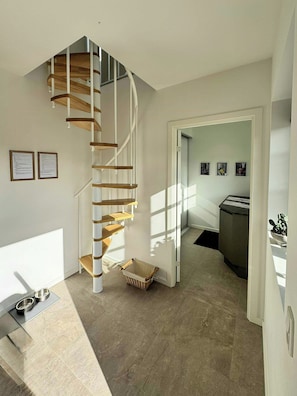 A modern interior with a spiral staircase, a dining area with a metal table and chairs, and a kitchen area visible through an open door.