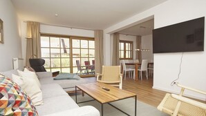 Ein modernes Wohnzimmer mit einer Couch, einem Couchtisch aus Holz, einem an der Wand montierten Fernseher und einem Essbereich mit Stühlen und Tisch.