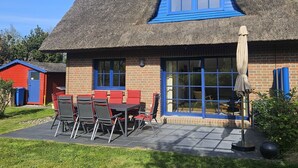 Ein Haus mit einem Strohdach, mit blauem Fenster und blauer Tür, einer Terrasse mit Tisch und Stühlen und einem kleinen Schuppen im Hintergrund.