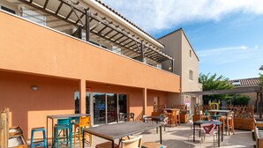 Uma área de pátio externa com mesas e cadeiras, um espaço coberto para sentar e um edifício com telhado inclinado.