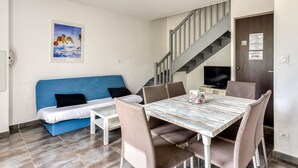 Quarto com sofá azul, mesa de jantar com cadeiras, televisão e escada.