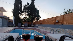 Ambiente junto à piscina com dois copos de vinho tinto, uma cesta de frutas e cadeiras de descanso.