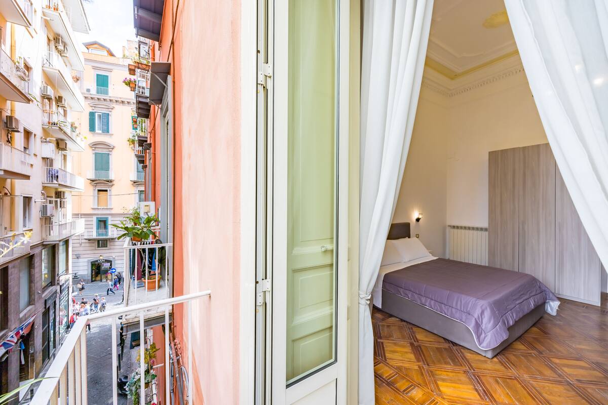 A balcony with a view of a narrow street, a bed with a purple bedspread, and a white curtain partially drawn.