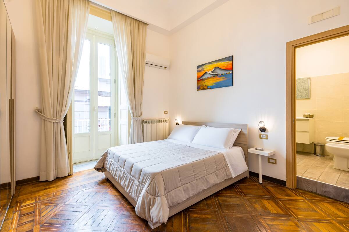 A hotel room with a bed, a nightstand, a painting, and a bathroom visible through an open door.