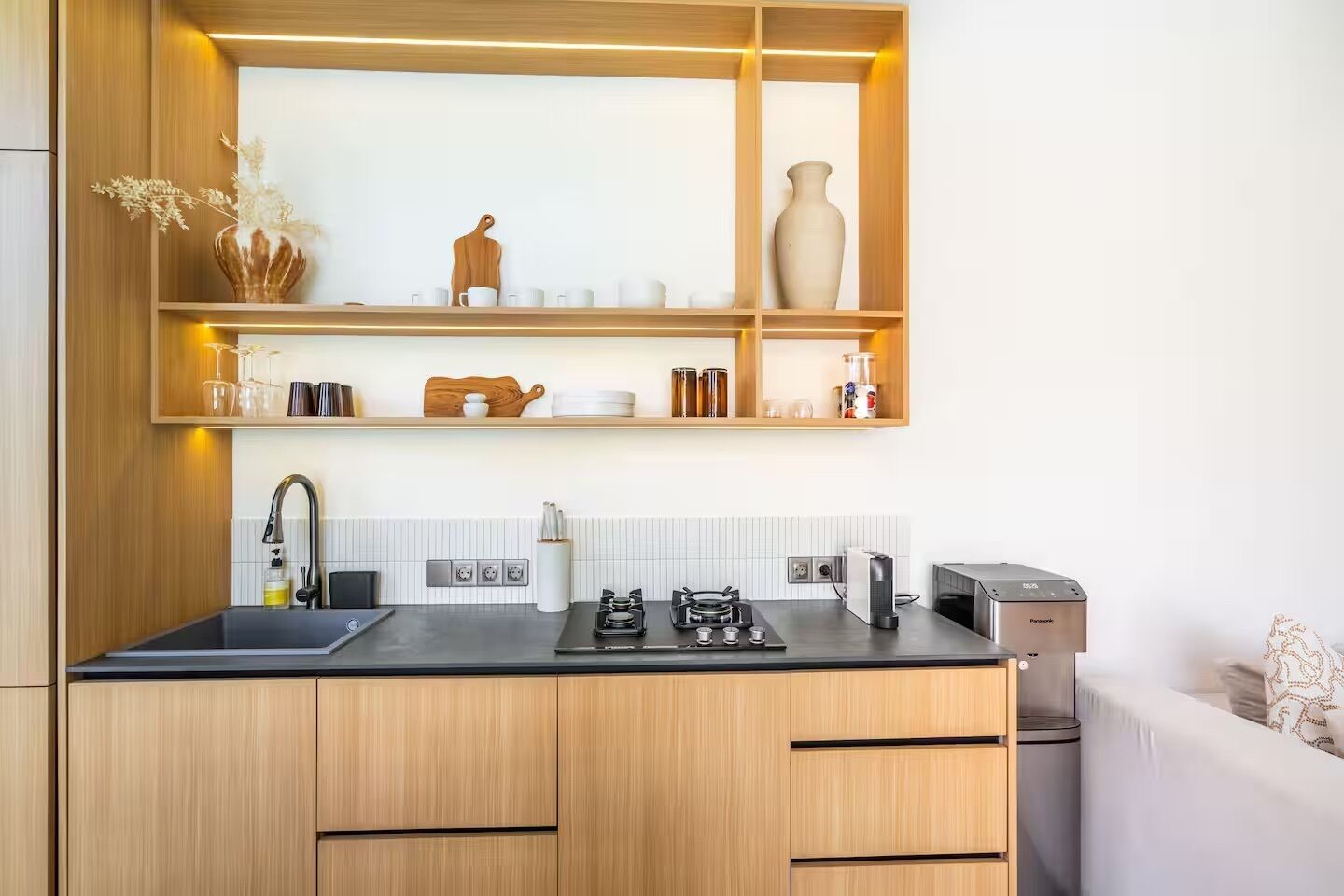 A modern kitchen with wooden cabinets, a black countertop, a gas stove, a coffee machine, and a built-in shelf with decorative items.
