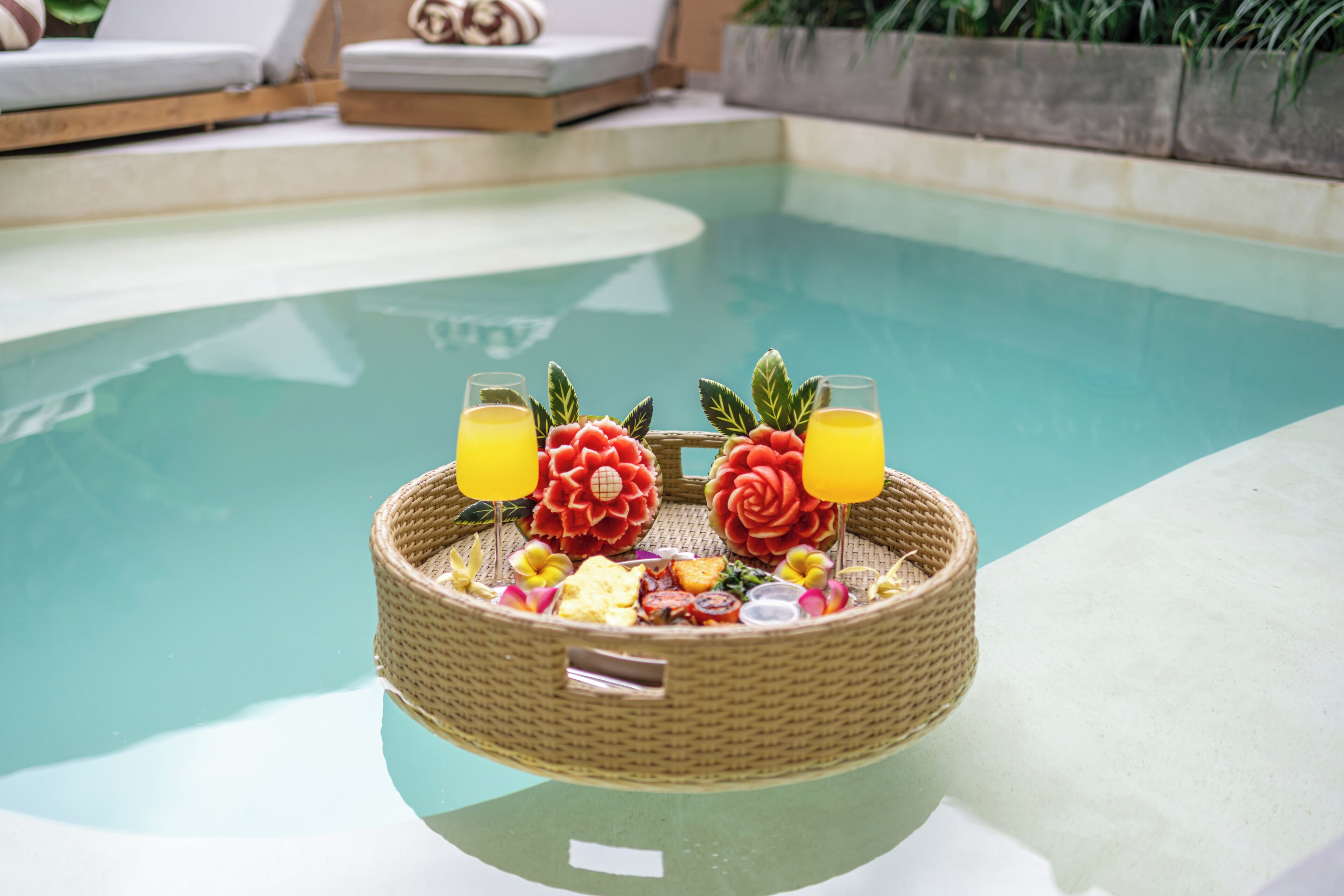 A poolside setting with a wicker basket containing food and drinks, a lounge chair, and a small table.