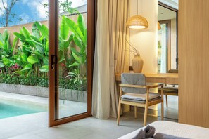 A hotel room with a glass door leading to a pool, a wooden desk, a chair, and a large window. - Berawa Week Villas (Canggu)