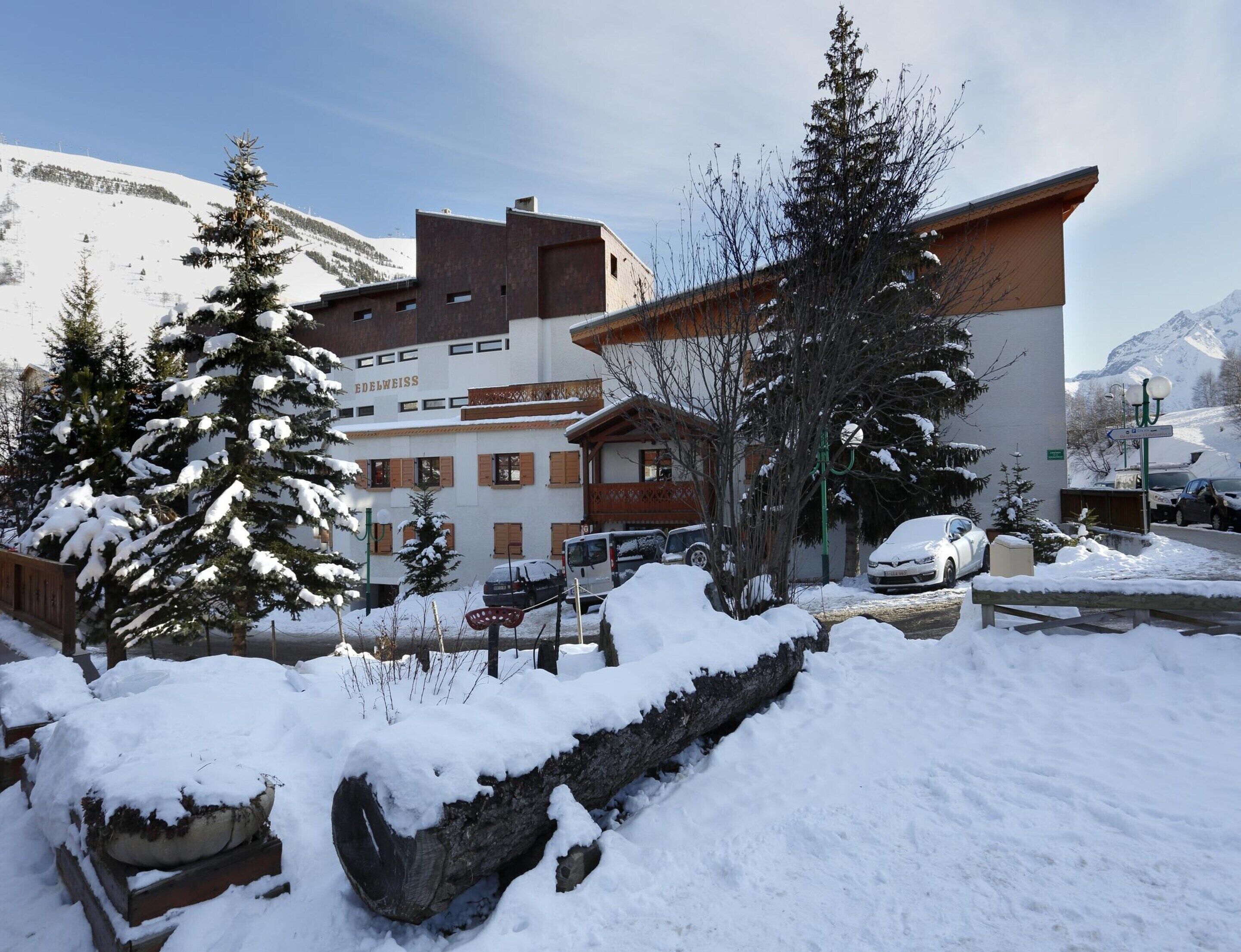 Un hotel con un paisaje nevado, una montaña al fondo y un estacionamiento con coches.