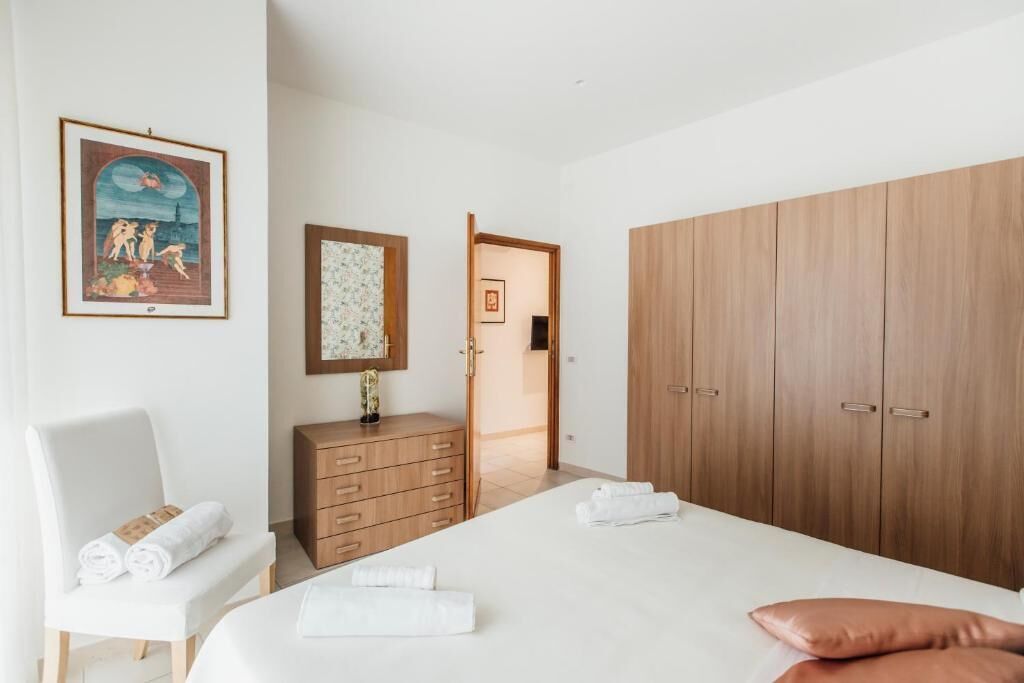 A hotel room with a bed, a wooden wardrobe, a bench with towels, a small cabinet, and a framed picture on the wall.