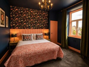 Ein Schlafzimmer mit blumigem Wandpapier, einem Bett mit gemustertem Überwurf, zwei Nachttischlampen, einer Chandelier und einem Fenster mit Vorhängen.