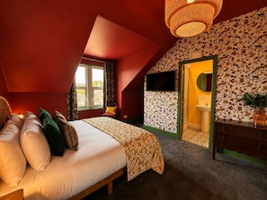 Ein Schlafzimmer mit Bett, Fenster, Fernseher und einem Badezimmer mit Waschbecken und Spiegel.