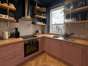 Eine moderne Küche mit rosa Schränken, einem schwarzen Herd, einer Kaffeemaschine und einem Fenster mit Blick auf Gebäude.