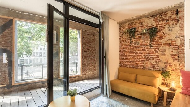 Una habitación con pared de ladrillo, sofá amarillo, una mesa con planta y ventana con vista a la calle.