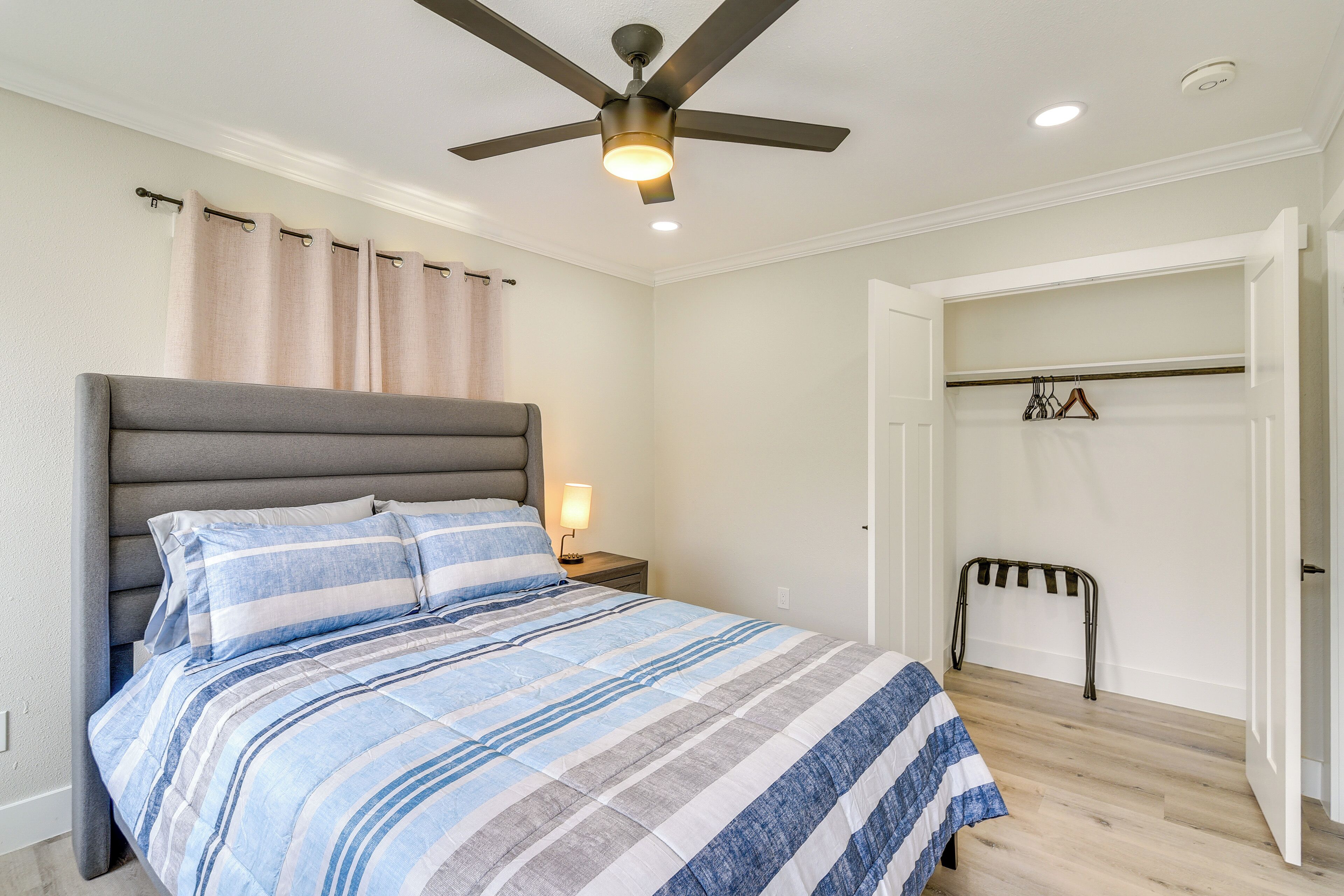 A bedroom with a bed, a ceiling fan, a nightstand, a lamp, a coat rack, and a door leading to another room.