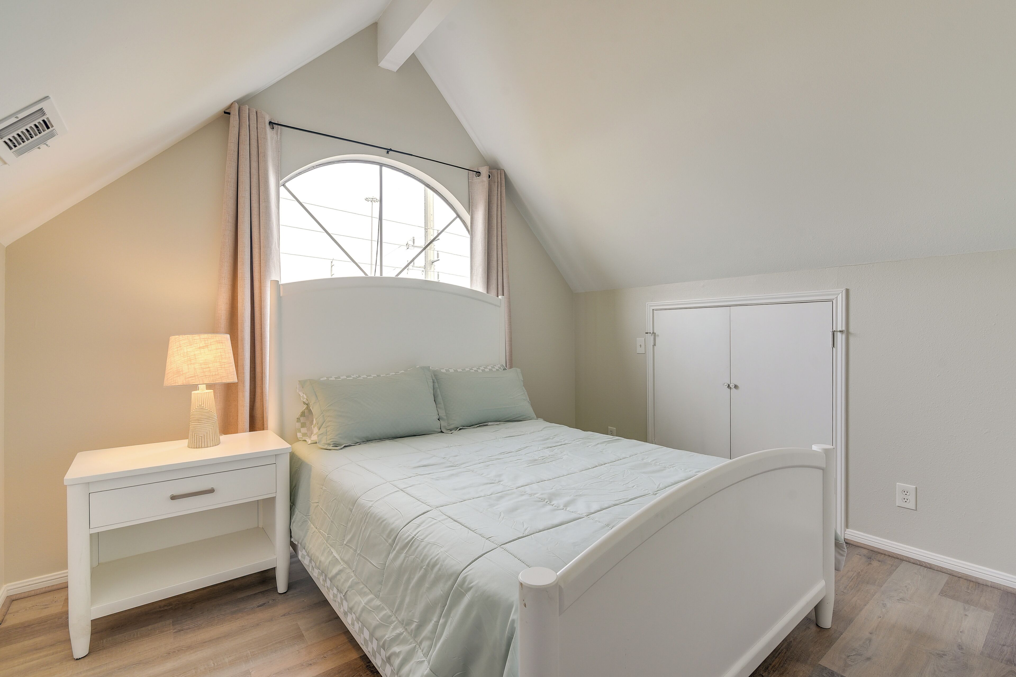 A bedroom with a bed, a nightstand, a lamp, a window with curtains, and a door.