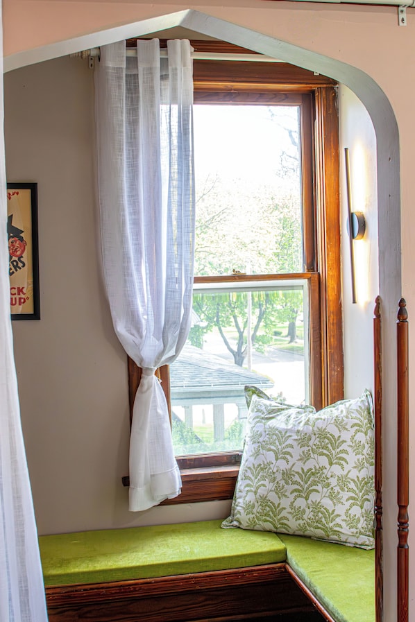 A room with a window, a bed with a green mattress, a white pillow with a green pattern, and a white curtain.