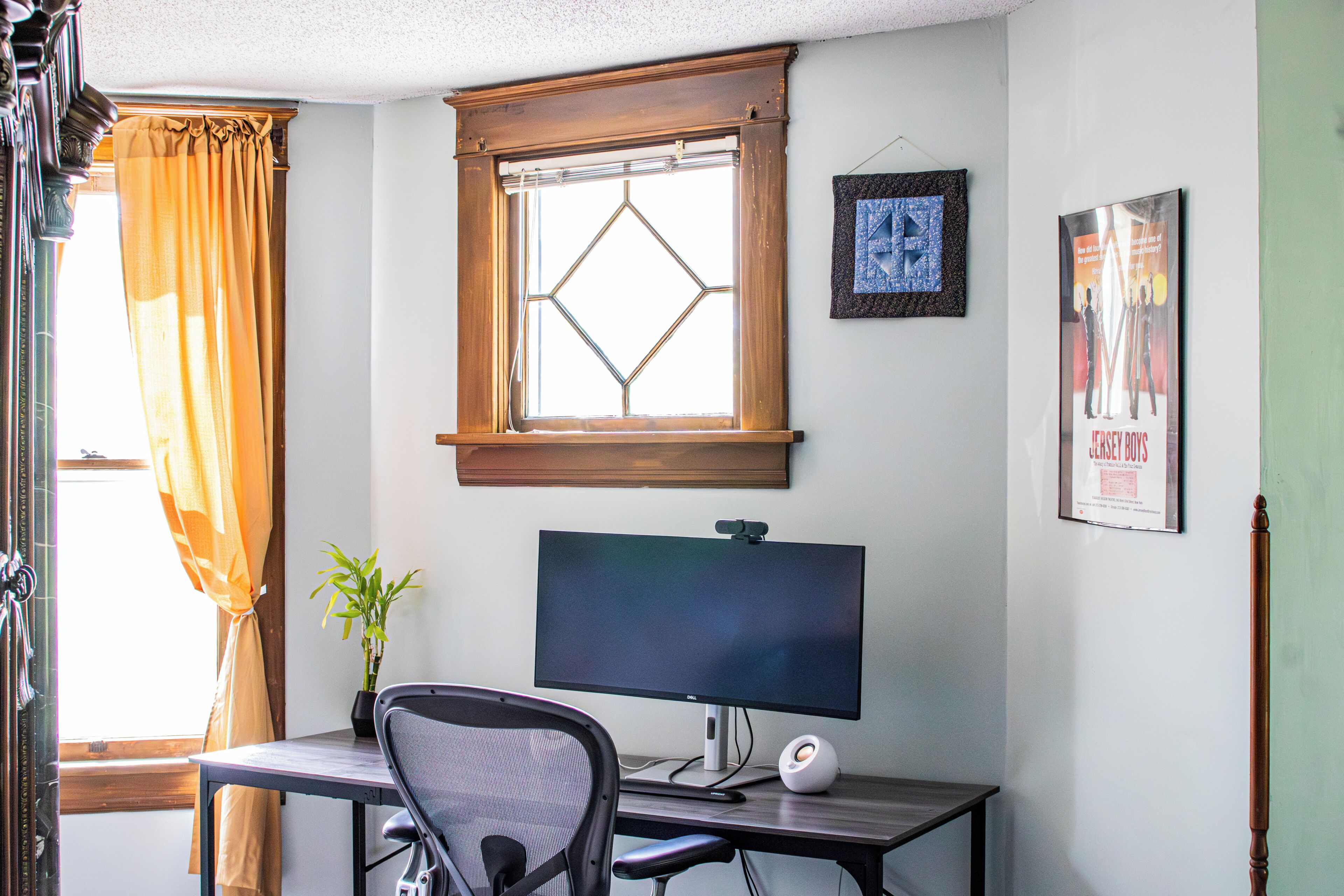 Een kamer met een bureau, een computer monitor, een stoel, een kamerplant, een raam met gordijnen, en een lijst aan de muur.