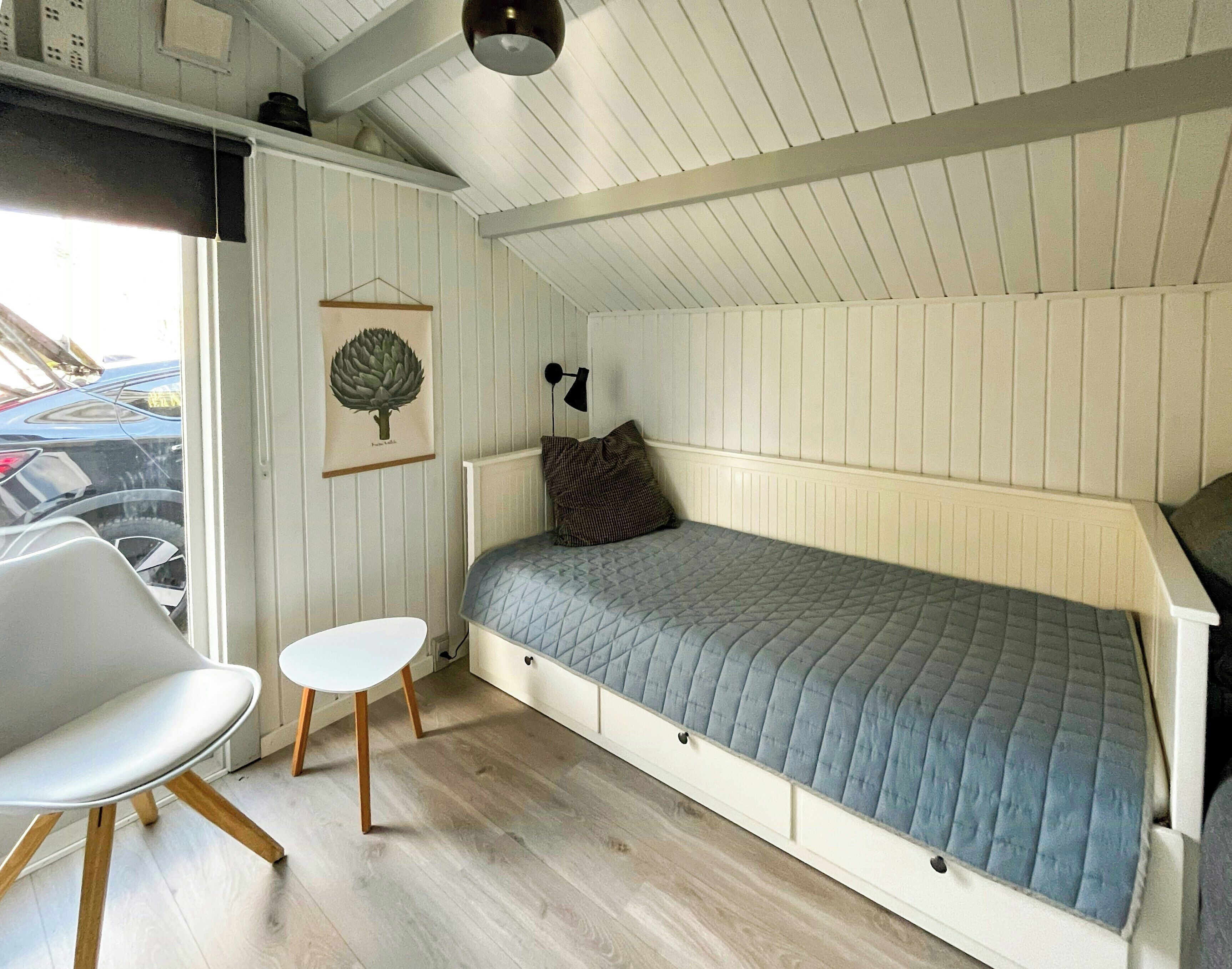 A small, single-bed room with a sloped ceiling, a white chair, a small table, and a wall-mounted lamp.