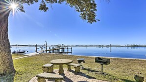 A picnic area with stone tables and benches, a grill, and a view of a calm lake with a dock in the background. - Dock Access! Condo 5 Mi to Okaloosa Island Beach (Mary Esther)