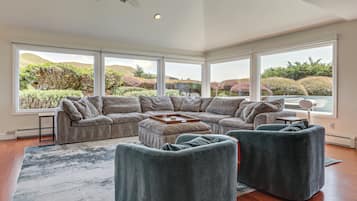 Ein modernes Wohnzimmer mit einem Ecksofa, zwei Sesseln, einem Couchtisch und großen Fenstern mit Blick auf eine angelegte Hügellandschaft.