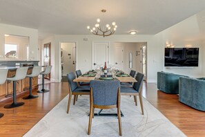 A dining area with a long wooden table, multiple chairs, a chandelier, and a large flat-screen TV mounted on the wall. - Dog-Friendly Haven w/ Stunning Views in Bodega Bay (Bodega Bay)