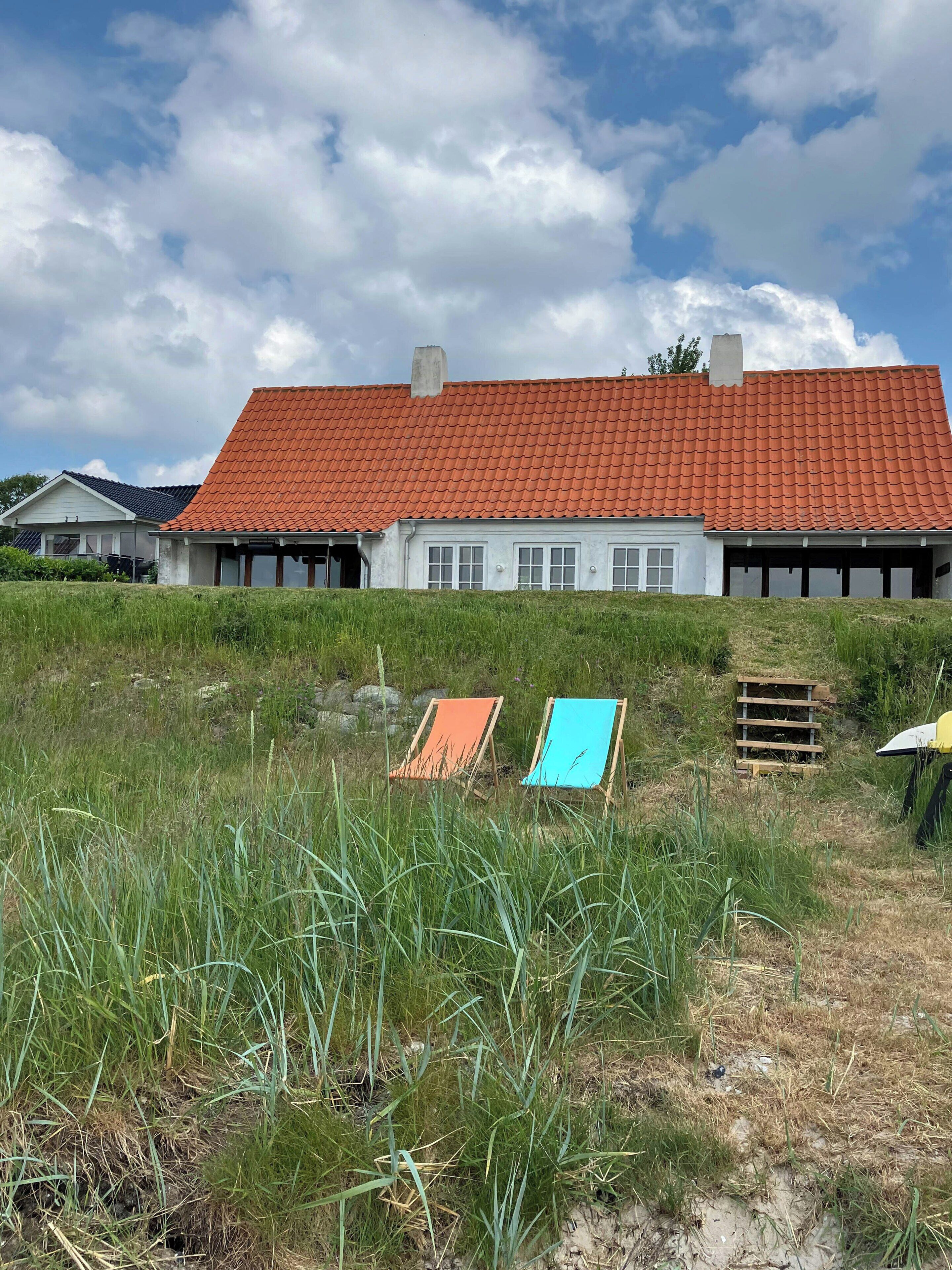 A house with a red-tiled roof, a garage, and a grassy foreground with two colorful chairs.