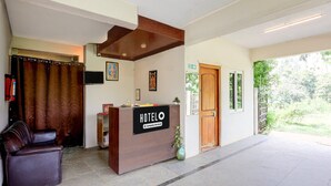 A hotel reception area with a wooden reception desk, a leather chair, a potted plant, and a door leading outside.