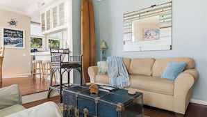 A living room with a beige sofa, a blue and brown surfboard, a vintage suitcase, and a wall with a striped mirror. - Ocean View Manhattan Beach Townhome | Steps to Beach (Manhattan Beach)