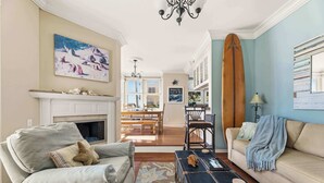 A living room with a fireplace, a dining area, and a surfboard. - Ocean View Manhattan Beach Townhome | Steps to Beach (Manhattan Beach)